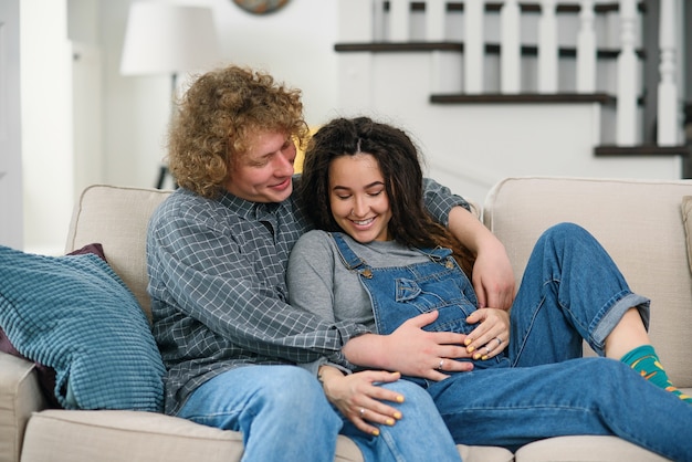 Marido com cabelos cacheados tocando a barriga de grávida de sua encantadora esposa alegre que estava deitada no sofá na aconchegante sala de estar