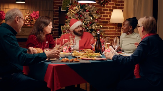 Marido divertido con traje de Santa Claus que ofrece regalos a la familia mientras celebra las vacaciones de invierno en casa. Hombre sano disfrazado de Santa Claus dando regalos familiares festivos en la cena de Navidad.