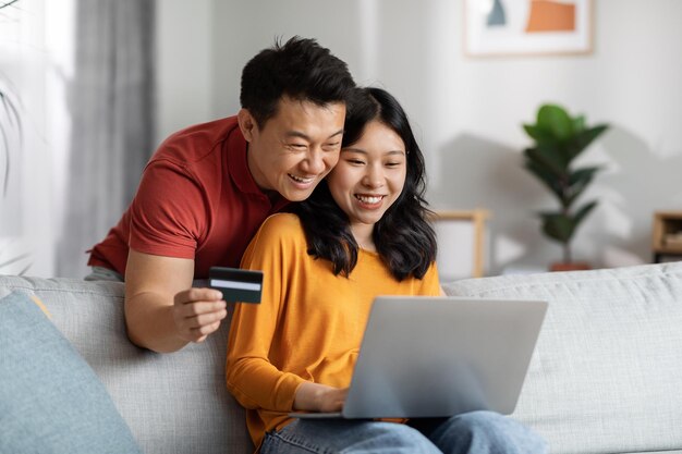 Marido e esposa asiáticos sorridentes usando computador e cartão de crédito