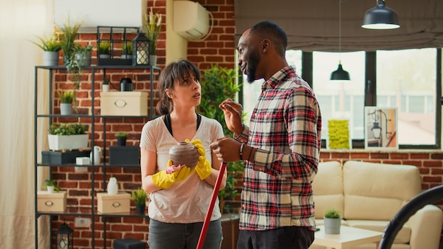 Marido e esposa diversificados limpando a casa com aspirador e panos, mulher usando limpador multiuso para varrer a sujeira das prateleiras. pessoa afro-americana aspirando pisos de madeira em casa.