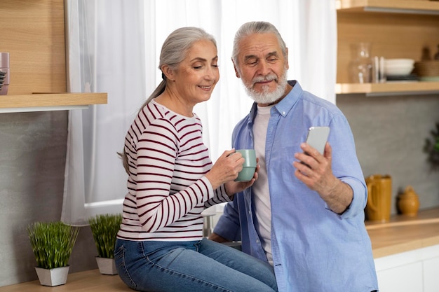 Marido e esposa idosos sorridentes relaxando na cozinha com smartphone e café