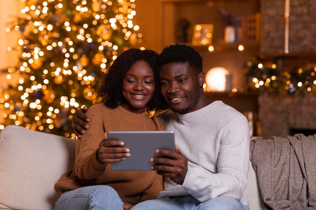 Marido e esposa negros milenares sorridentes fazem videochamada no tablet, assistem ao vídeo, sentam-se no sofá na sala de estar