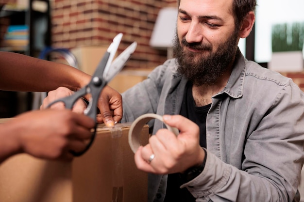 Marido e mulher cortando fita adesiva com tesoura para mover em apartamento alugado, usando rolo adesivo para embalar móveis para transporte de armazenamento. Dia de mudança para novos começos. Fechar-se.