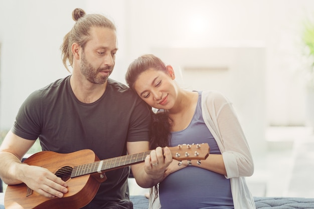 Marido con esposa embarazada quedarse en casa encantadora tocando música de guitarra para besar al bebé y mostrar amor juntos