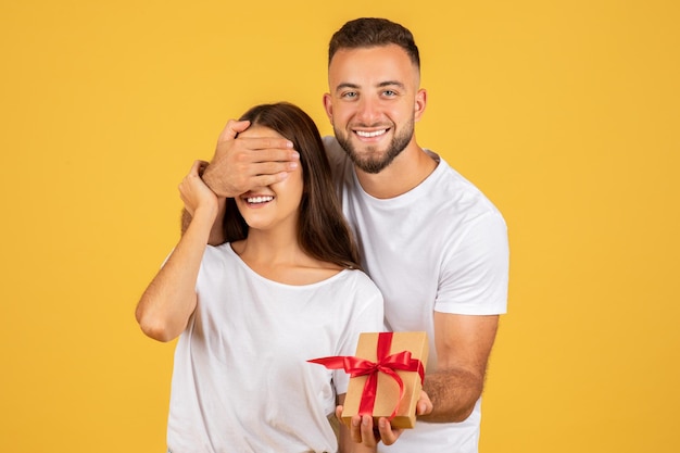 Foto el marido europeo del milenio positivo cierra los ojos a la esposa y le da una caja de regalo.