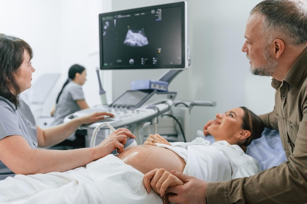 Foto el marido feliz está sentado y apoyando a su esposa la mujer embarazada está acostada en el hospital el médico hace ultrasonido