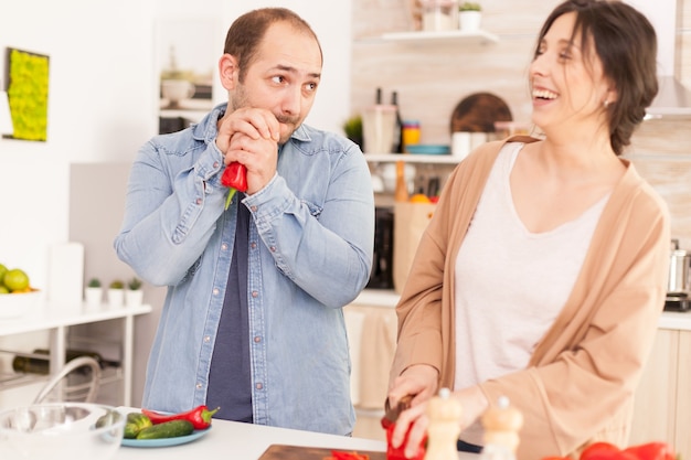 Marido mirando a la esposa mientras ella sonríe a causa de él en la cocina. Cortar pimiento rojo en picadora de madera. Divertido feliz pareja de enamorados en casa pasar tiempo juntos, cocinar sano y sonriente