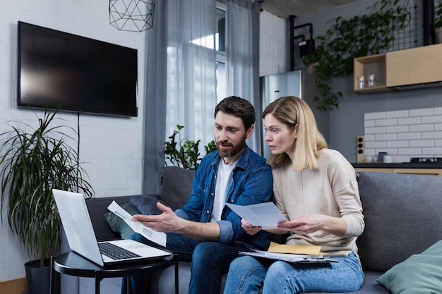 Marido y mujer familia joven trabajando juntos en las finanzas del hogar discutiendo facturas usando una computadora portátil sentada en un sofá en casa