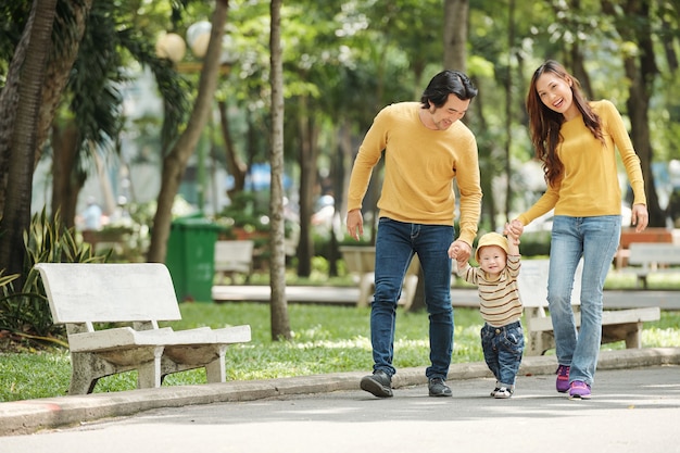 Marido, mulher e bebê vietnamitas alegres de jeans e suéteres andando no parque local em um dia ensolarado