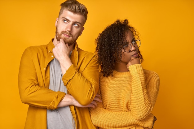 Marido ruivo e esposa afro-americana perplexos segurando o queixo e franzindo os lábios olham com hesitação