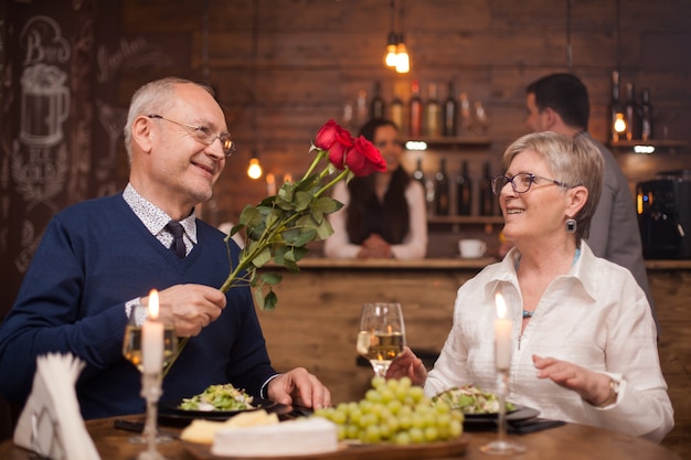 Marido de unos sesenta años regalando rosas a su esposa en un restaurante. Feliz esposa. Ancianos sonriendo. Hombre y mujer maduros.