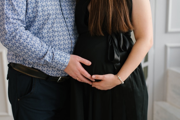 El marido sostiene su mano sobre el vientre de una mujer embarazada en un vestido negro de cerca. futuros padres que esperan un bebé. cuidados y maternidad