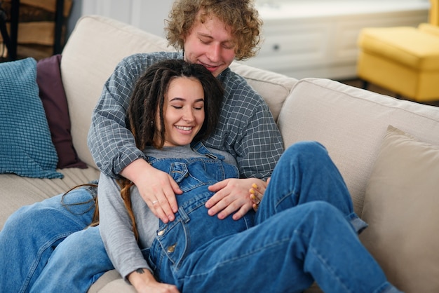 Marido tocando a barriga de grávida de sua encantadora esposa alegre que estava deitada no sofá em uma casa aconchegante