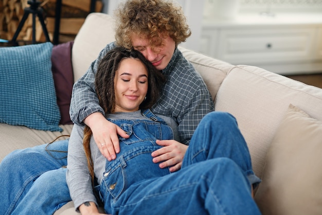 Marido tocando a barriga de grávida de sua encantadora esposa alegre que estava deitada no sofá em uma casa aconchegante