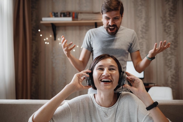 Marido zangado gritando com a esposa sentada no sofá e cobrindo os ouvidos com fones de ouvido