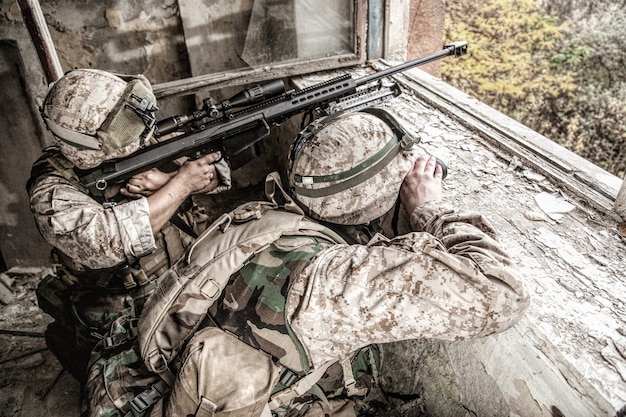 Marines Scharfschützenteam, bewaffnet mit großkalibrigem, anti-materiellem Scharfschützengewehr, das sich in einem zerstörten Stadtgebäude versteckt, feindliche Ziele aus der Entfernung aus dem Unterstand abschießt und im Hinterhalt sitzt Militärisches Feuergefecht in der Stadt