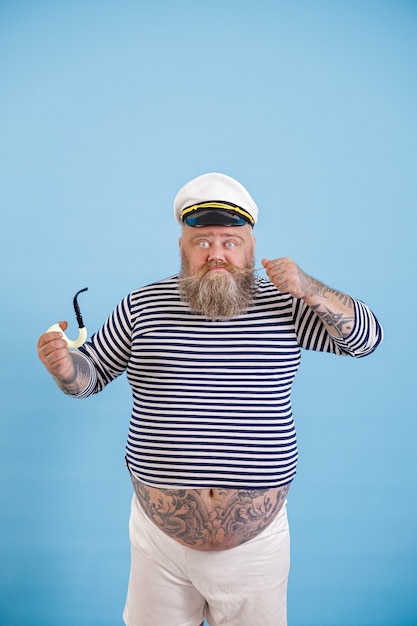 Marinheiro obeso barbudo com cachimbo de fumar aperta bigode em fundo azul claro