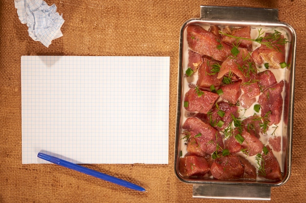 Mariniertes Fleisch in Milch und Gemüse in einer tiefen Stahlschale auf einem selbstgemachten Tuch mit sauberen Blättern Papier und einem Kugelschreiber