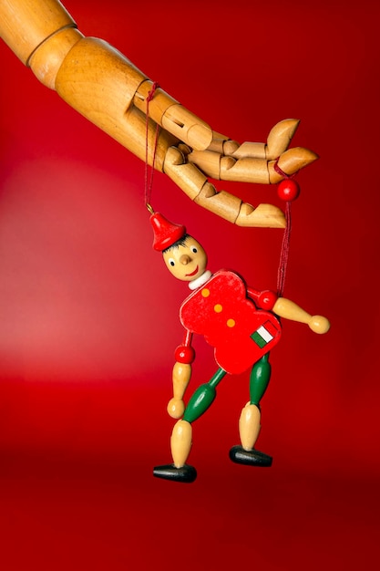 Una marioneta controlada por una mano de madera sobre un fondo rojo.