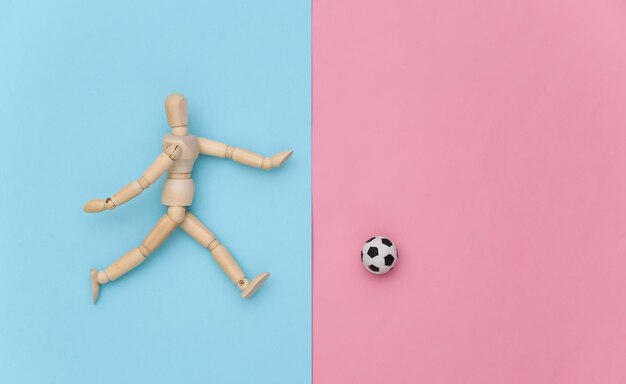 Marioneta de madera jugando al fútbol con una pelota sobre fondo azul rosa