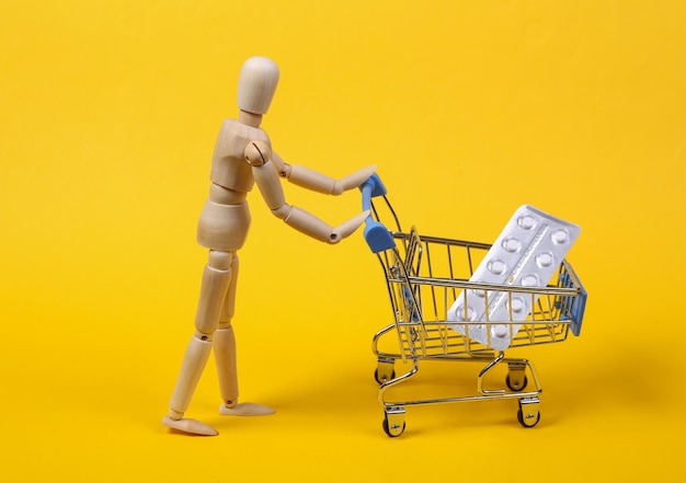 La marioneta de madera sostiene un carrito de compras con una pastilla de ampolla sobre fondo amarillo. concepto de medicina