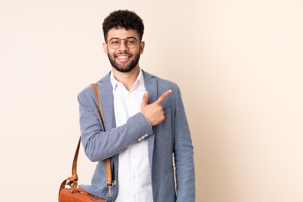 Marokkanischer Mann des jungen Geschäfts lokalisiert auf beige Wand, die zur Seite zeigt, um ein Produkt zu präsentieren