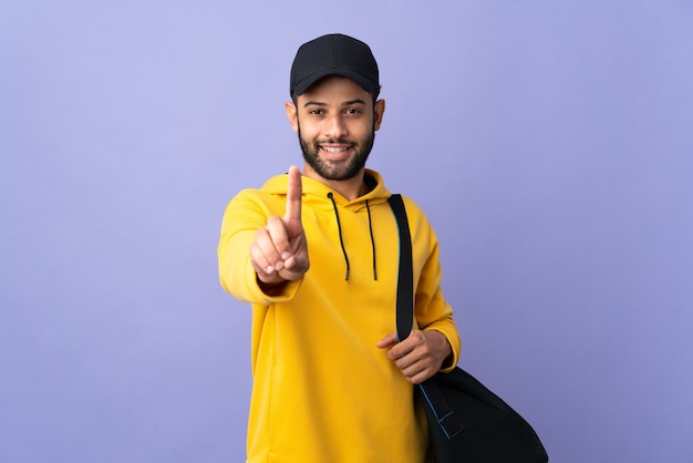 Marokkanischer Mann des jungen Sports mit Sporttasche lokalisiert auf lila Wand, die einen Finger zeigt und hebt