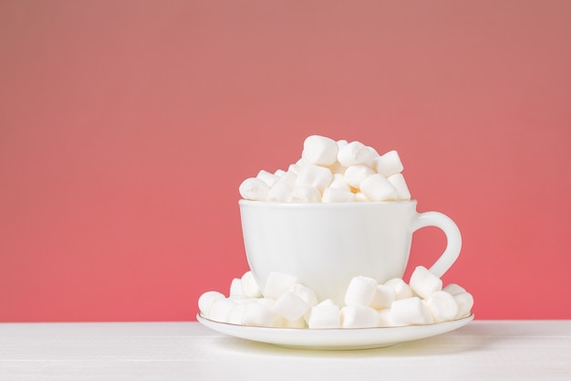 Marshmallow em uma xícara branca e pires sobre uma mesa branca sobre um fundo vermelho. Sobremesa deliciosa.
