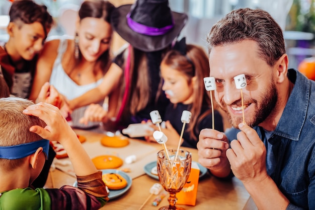 Marshmallows em palitos. Pai barbudo sorridente segurando marshmallows em palitos enquanto celebra o Halloween com esposa e filhos