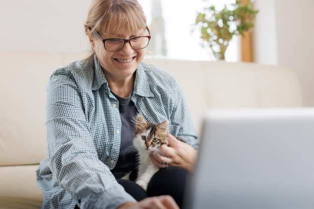 Marure Frau in blauem Hemd sitzt mit einer Katze auf dem Schoß am Holztisch zu Hause mit Laptop und Notebook und arbeitet oder kauft online ein