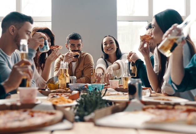 Más risa con amigos. Grupo de jóvenes en ropa casual comiendo pizza y sonriendo mientras tienen una cena en el interior