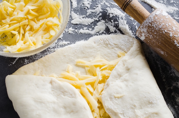 Masa enrollada, harina para hornear, queso rallado y rodillo sobre una pizarra oscura.