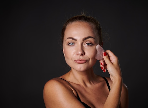 Masaje facial guasha con piedra de jade Mujer rubia de mediana edad con piel bronceada masajeando la piel facial con procedimiento de spa de automasaje con raspador de belleza guasha en casa