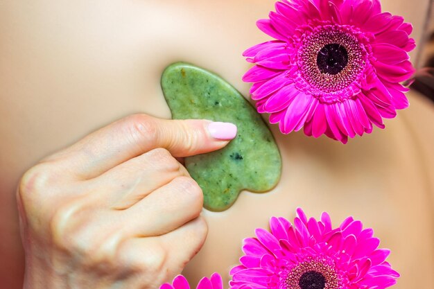 Foto masaje facial de guasha con piedra verde jade en un primer plano de la mano de una mujer con una flor