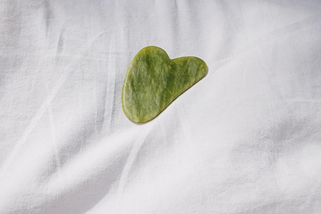 Masajeador gua sha de piedra de jade verde sobre un fondo blanco mínimo con sombra en un día soleado de verano Rutina matutina de cuidado de la piel de productos de belleza Paquete orgánico y de bienestar Concepto de spa