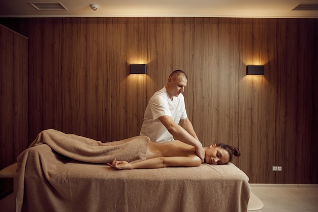 Masajista masculino mimando el cuello a la mujer joven en toalla, masaje profesional. Masaje y relajación, cuidado corporal y cutáneo. Señora atractiva en el salón de spa