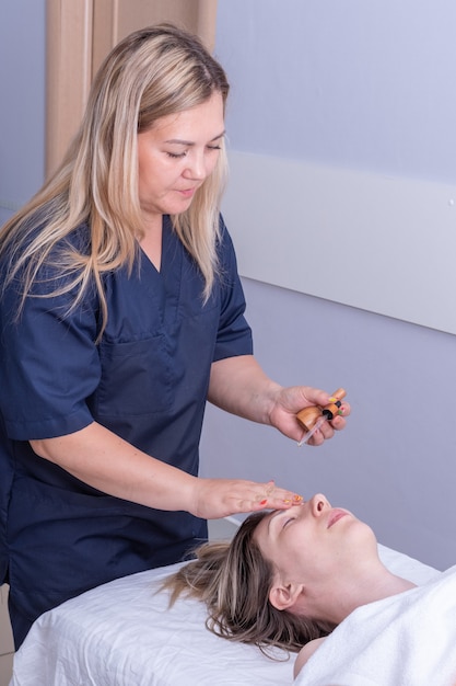 Masajista niña gotea aceite esencial con una pipeta en la cara de la niña para masajear la cara y el cuello, marco vertical. Masaje anti-edad de rostro y cuello. Cuidado cosmético para la piel del rostro y el cuello.