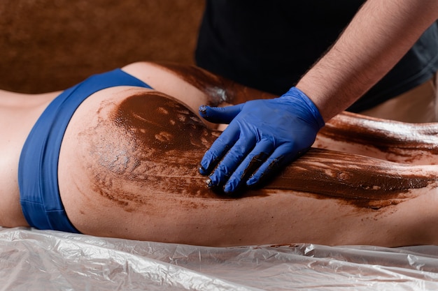 El masajista unta chocolate en las piernas y muslos de la niña en el centro de spa. Envoltura de chocolate de procedimiento de belleza.