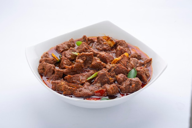 Masala de carne ou assado, receita caseira estilo kerala guarnecida com pedaços de coco e folhas de curry que é disposta em tigela branca com fundo branco.