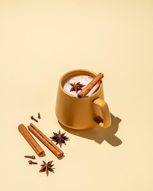 Masala latte tea Bebida tradicional india con una mezcla de especias y hierbas Una taza amarilla con una bebida espumosa rodeada de palitos de canela y anís estrella sobre un fondo amarillo