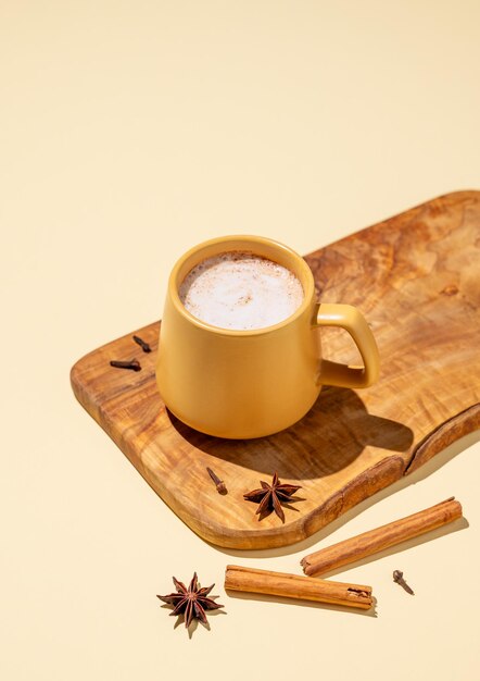 Foto masala latte tee traditionelles indisches getränk mit einer mischung aus gewürzen und kräutern ein gelber becher mit einem schaumigen getränk, umgeben von zimtstäbchen und sternanis auf einem holzbrett auf einem gelben hintergrund