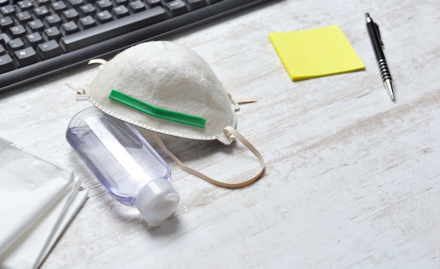 Máscara antipolvo para proteger la cara y una botella de desinfectante para manos en un escritorio junto al teclado y papel de notas amarillo con un bolígrafo