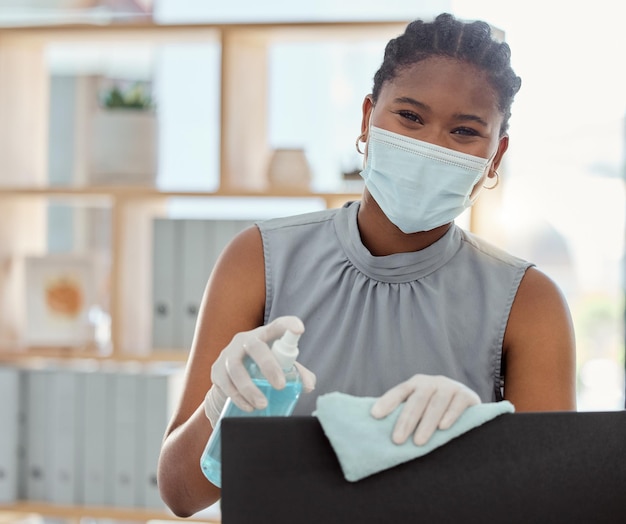 Máscara Covid e desinfetante com uma mulher negra de negócios limpando seu computador e equipamento de escritório no trabalho Retrato higienizando e limpando com uma funcionária durante a pandemia do vírus corona