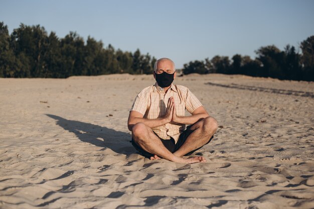 Máscara de medicina vestindo homem sênior em pose de lótus, sentado na areia. Conceito de calma e meditação.