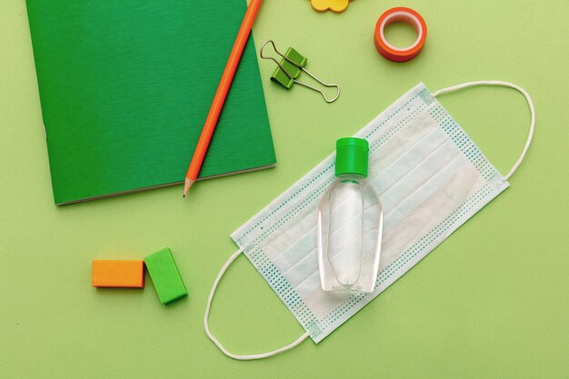 Foto máscara de proteção médica, gel de desinfetante de mãos e suprimentos escolares em fundo verde pastel, vista superior de volta aos dias de coronavírus na escola