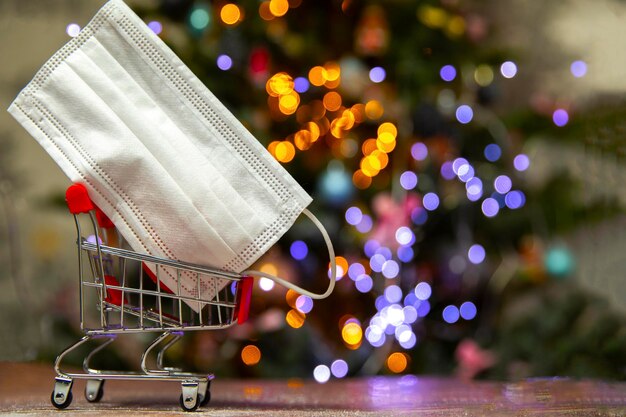 Máscara de segurança protetora em uma cesta de compras de ferro perto da árvore de Natal, Covid-19, coronavírus, conceito de férias com espaço de cópia e fundo de luzes bokeh