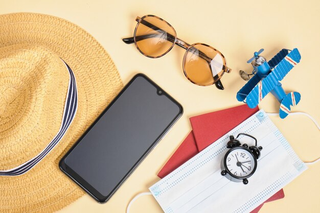 Máscara facial, chapéu de palha, smartphone, óculos de sol, despertador e passaporte em fundo bege, viagem durante o bloqueio, conceito de férias na praia segura