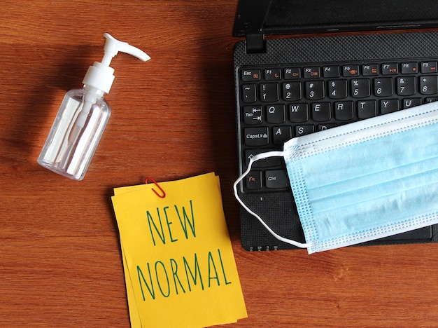 Foto máscara facial de desinfetante para as mãos para laptop e papel escrito com texto novo normal em fundo de madeira