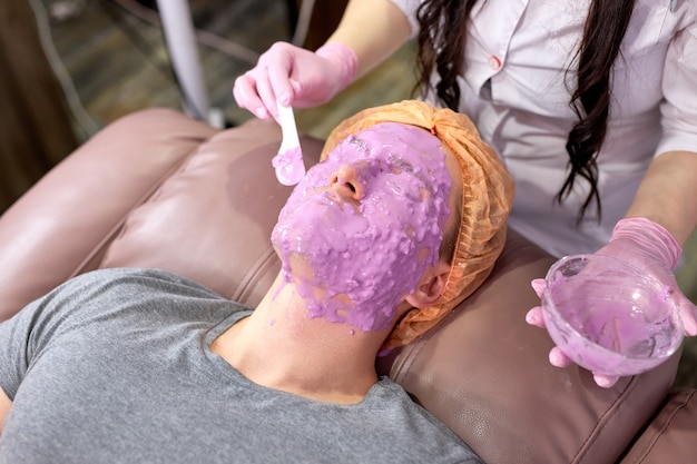 Máscara facial de hombre en salón de spa, procedimiento de belleza masculino. masaje en la cara. Esteticista recortada con recipiente aplicando líquido sobre la piel, tratamiento facial contra el acné