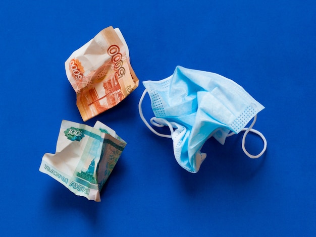Máscara médica amassada contra coronovírus e dinheiro russo amassado sobre um fundo azul.